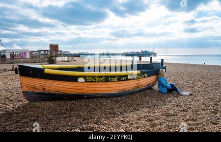 Brighton UK 6th March 2021 - Zeit, um in der Sonne für diese Dame am Brighton Strand gegen ein altes Fischerboot an einem sonnigen, aber kühlen Morgen entlang der Südküste zu sitzen : Credit Simon Dack / Alamy Live News Stockfoto