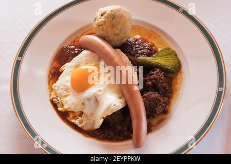 Fiakergulasch oder Herrengulasch Wiener Rindfleisch Gulasch garniert mit einem Brotknödel, Frankfurter Wurst, gebratene Ei und Gherkin auf einem Teller Stockfoto