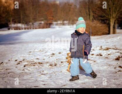 POZNAN, POLEN - 17. Jan 2021: Kleiner drei Jahre alter polnischer kaukasischer Junge in warmen Kleidern, der einen hölzernen Schlitten mit einer Linie auf Schnee bei einem pa zieht Stockfoto