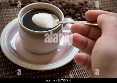 Ein Mann Hand hinzufügen weißen Zucker mit metallischen Löffel über Eine Tasse voll Kaffee auf dem Teller auf einem Bambus Mat.Kaffeestube Stockfoto