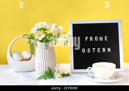 Text Frohe Ostern bedeutet Frohe Ostern. Text auf Briefbrett mit Freesie Blumen auf Tisch. Schwarzer Briefbogen, Text. Natürliche Ostereier, Teetassen auf Stockfoto