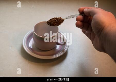 Die Zubereitung von löslichem Kaffee. Gefriergetrockneter Kaffee in einem Löffel über einer Tasse auf dem Teller. Die Hand eines Mannes, der Kaffee macht. Stockfoto