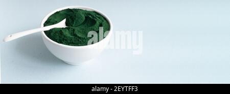 Banner Spirulina Pulver in einer weißen Schüssel mit einem Teelöffel auf einem blauen Hintergrund mit Platz für Text. Gesunde Ernährung Konzept. Horizontale Ausrichtung. Stockfoto