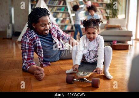 Ein junger Vater schiebt seine kleine Tochter in heiterer Atmosphäre zu Hause auf das Skateboard. Familie, zusammen, Liebe, Spielzeit Stockfoto