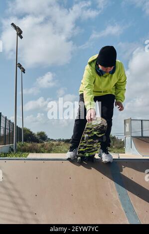 Jung, Teenager, mit einem Skateboard, halten das Board mit der Hand, um den Hang hinunter zu starten, auf einer Strecke, Skateboarding, Tragen von Kopfhörern, grün sw Stockfoto