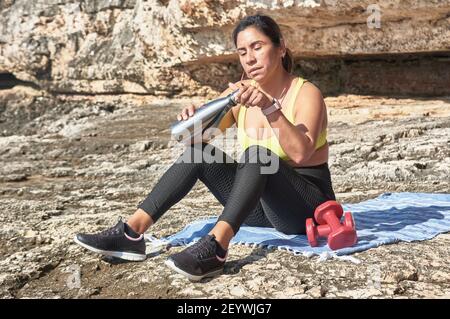 Lateinische Frau, mittleren Alters, ausruhen, Kraft zurückgewinnen, essen, Trinkwasser, nach einem Fitness-Studio-Sitzung, Kalorien zu verbrennen, fit zu halten, im Freien am Meer Stockfoto