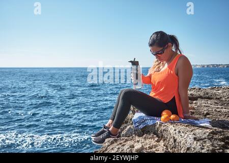 Lateinische Frau, mittleren Alters, ausruhen, Kraft zurückgewinnen, essen, Trinkwasser, nach einem Fitness-Studio-Sitzung, Kalorien zu verbrennen, fit zu halten, im Freien am Meer Stockfoto