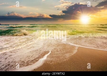 Meeresflut auf einem bewölkten Sonnenaufgang. Grüne Wellen krachenden goldenen Sandstrand im Morgenlicht. Sturmwetter nähert sich. Sommerferienkonzept Stockfoto