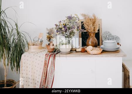 Startseite Küche rustikale Inneneinrichtung der Wohnung mit Tisch, Ton und Holz Utensil, Blumen, Heimpflanzen, Textil. Stilvolles Wohndekor im offenen Raum. Stockfoto