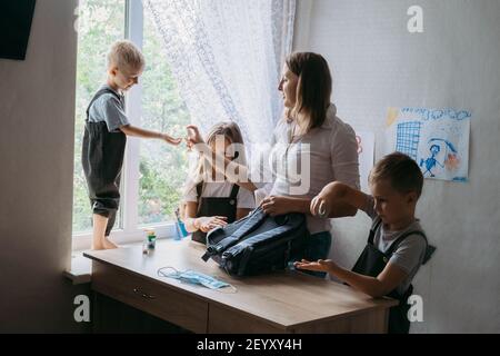 Zurück zur Schule in neuer Normalität. Mutter hilft Schulkindern, ihre Rucksäcke für die Schule zu packen und gibt ihnen Gesichtsmasken und Desinfektionsmittel. Medizinische Maske zu Stockfoto