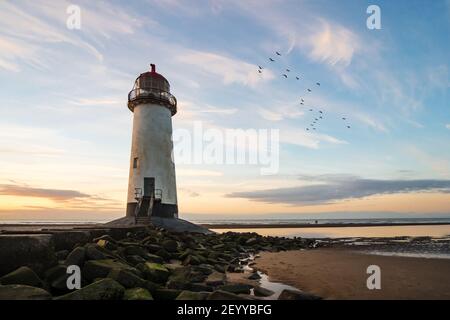 Wunderschöner Leuchtturm am Meer, goldener Sonnenuntergang mit Felsen, die zu Treppen führen, wenn die Meeresströmung hereinkommt. Vögel fliegen zum Horizont Sonne reflektierendes Wasser Stockfoto