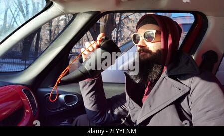 Nahaufnahme eines bärtigen jungen Mannes, der Kaffee zum Mitnehmen trinkt. Ein bärtiger Mann mit Schnurrbart schaut aus dem Fenster im Auto. Mann im Auto Stockfoto