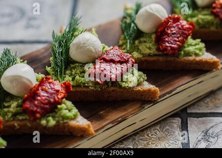 Mini-Dreieck Canapes Bruschetta mit Avocado, Mozarella und getrockneten Tomaten auf dem rustikalen Brett auf dem vintage gekachelten Hintergrund Stockfoto