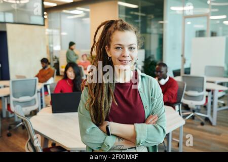 Junge lächelnde Geschäftsfrau oder Designerin in Casualwear, die ihre Arme kreuzt Bei der Arbeit vor der Kamera stehen Umwelt Stockfoto