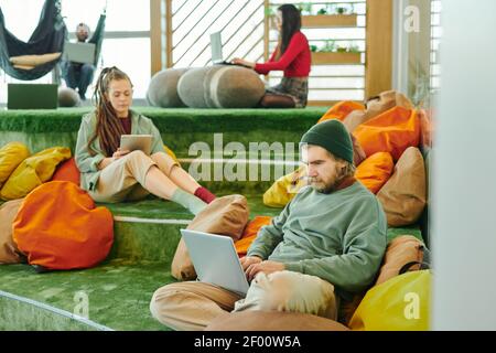 Mehrere junge interkulturelle Manager von großen modernen Open Space Büro Mit Laptops, während Sie auf grünem Boden gegen Kissen sitzen Stockfoto