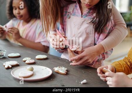 Hände einer jungen Frau, die ihrer kleinen Tochter mit Verglasung hilft Frische hausgemachte Kekse oder Lebkuchen, während beide am Tisch stehen Stockfoto