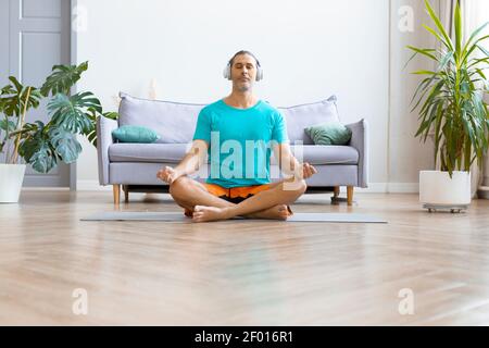Foto eines Mannes mittleren Alters, der zu Hause Yoga praktiziert. Er sitzt in der Lotusposition mit Kopfhörern und hört Meditationsmusik. Workout zu Hause. Stockfoto