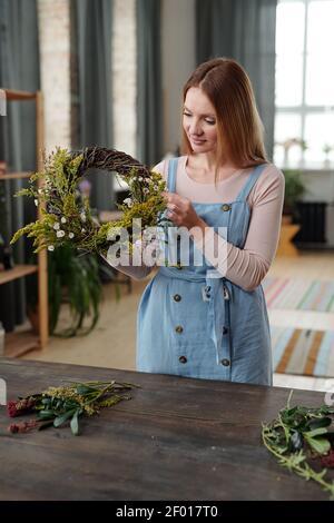 Glückliche junge blonde Frau in Casualwear stehen am Holztisch In häuslicher Umgebung und Vorbereitung Osterkranz für den Urlaub Stockfoto