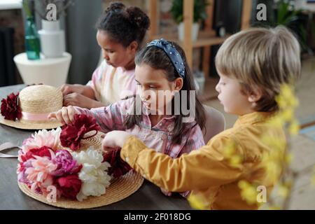 Gruppe von niedlichen interkulturellen Kinder in Casualwear sitzen am Tisch zu Hause und setzen handgemachte Blumen von weißen, rosa und karmesinroten Farben auf Hut Stockfoto