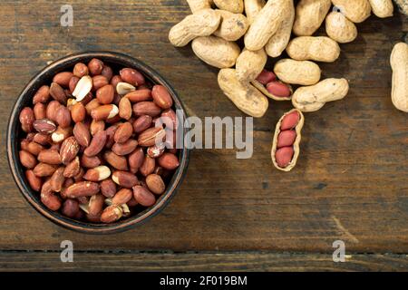 Geschälte Erdnüsse in einer Tonschüssel und auf einem Holztisch ungeschält. Draufsicht. Leerzeichen für Text. Stockfoto