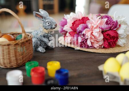 Graues Spielzeug Kaninchen, Korb mit bemalten Ostereiern und Hut mit handgefertigten weißen, rosa und karmesinroten Blumen auf Holztisch vor der Kamera Stockfoto