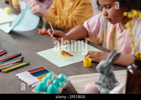 Cute serious Mixed-Race kleine Mädchen Malerei große Osterei auf Leeres Papier beim Sitzen am Holztisch auf dem Hintergrund Ihre beiden Freunde Stockfoto