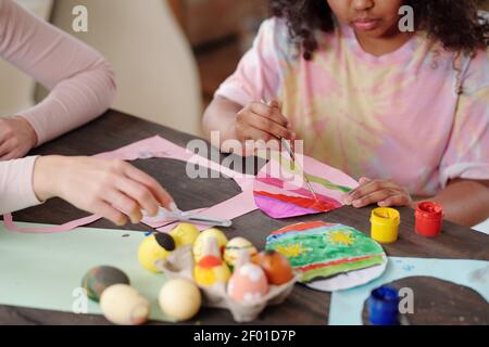 Serious Mixed-Race kleines Mädchen mit Pinsel Malerei Papier Osterei Beim Sitzen am Holztisch neben einer jungen Frau, die hilft Sie Stockfoto