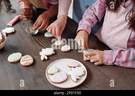 Hände von interkulturellen kleinen Mädchen und junge Frau in Casualwear Stehen am Holztisch und dekorieren hausgemachte Kekse mit Streuseln Stockfoto