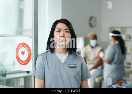 Junge zeitgenössische Klinikerin in Uniform vor Kamera gegen ihre Kollegin, die einen ihrer Patienten impft Stockfoto