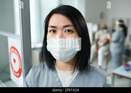 Junge Brünette weibliche Klinikerin in schützende Arbeitskleidung stehen vor Der Kamera, während ihre Kollegin die Patientin auf dem Hintergrund impft Stockfoto