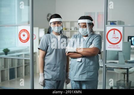 Zwei junge interkulturelle Klinikerinnen in schützender Arbeitskleidung im Stehen Vorderseite der Kamera gegen medizinische Büroinnenraum in modernen Kliniken Stockfoto