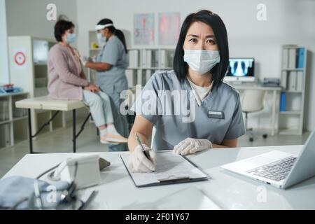 Junge asiatische Kliniker in schützende Arbeitskleidung sitzen am Schreibtisch und Ausfüllen des Dokuments, während die Kollegin die Patientin im Hintergrund impft Stockfoto