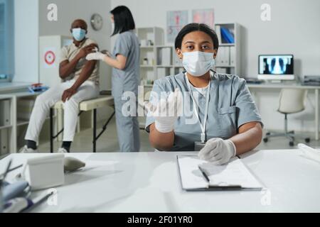 Junge Mixed-Race-Kliniker in schützende Arbeitskleidung Blick auf Sie während Medizinische Beratung während ihr Kollege geimpfte Patienten Stockfoto