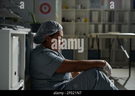 Seitenansicht der jungen müden gemischten Ärztin in Uniform Auf dem Boden sitzen und sich nach einem anstrengenden Arbeitstag entspannen In modernen Kliniken Stockfoto