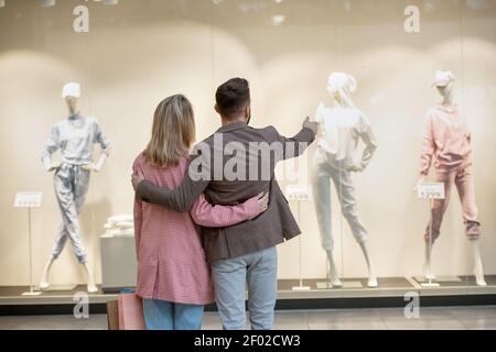 Rückansicht des jungen Paares, das vor dem steht Schaufenster und diskutieren Kleidung auf Schaufensterpuppen zusammen in der einkaufszentrum Stockfoto