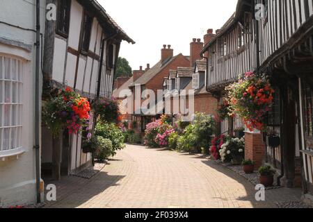 Schwarz-weiße Fachwerkhäuser mit hängenden Körben und Blumenblüten im Sommer in Shakespeare. Malz Mill Lane, Alcester, Warwick Stockfoto