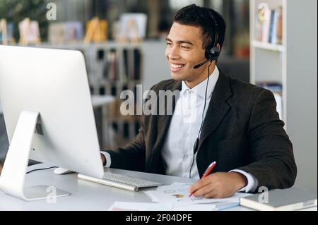 Zufriedener hispanischer männlicher Mitarbeiter auf Videokonferenz online. Glücklicher Geschäftsmann, der ein Headset trägt, auf den Laptop-Bildschirm schaut, Notizen macht, mit Kollegen kommuniziert, lächelt Stockfoto