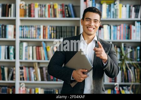 Portrait eines gutaussehenden hispanischen Geschäftsmannes oder Studenten in formell stilvollem Anzug, hält einen Laptop in den Händen und blickt mit freundlichem Lächeln direkt auf die Kamera Stockfoto