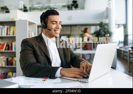 Freundlicher Geschäftsmann oder Freiberufler im Headset, kommuniziert per Videoanruf. Attraktive männliche Manager oder Berater sitzen am Schreibtisch in modernen Büro nutzt Laptop, chatten online Stockfoto