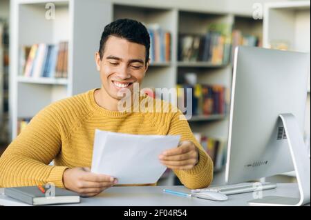 Fröhlicher männlicher Angestellter sitzt am Tisch im modernen Büro und lächelt. Hispanic Geschäftsmann macht wichtige Papiere, genießen eigene erfolgreiche Ergebnisse Stockfoto
