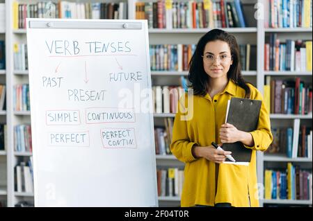 Selbstbewusste junge Tutorin Frau in Brille steht drinnen in der Nähe Flipchart. Wunderschöne Frau in stilvoller Kleidung sieht auf die Kamera, lächelt, arbeitet als Lehrer Stockfoto