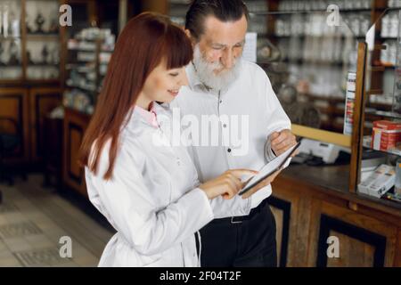 Junge professionelle junge Dame Apotheker hält Tablette und konsultiert bärtigen älteren Mann Kunden in alten alten alten Drogerie. Apotheker und Patient bei Stockfoto