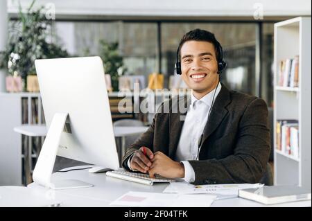 Portrait eines gutaussehenden hispanischen Angestellten in formellem Anzug und Headset schaut lächelnd auf die Kamera. Erfolgreicher Mann sitzt im modernen Büro, Beratung eines Kunden, Support-Service-Konzept Stockfoto