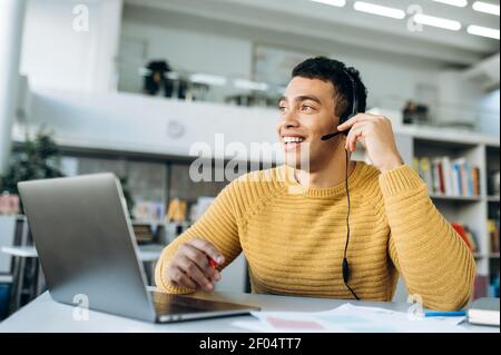 Fröhliche junge Erwachsene Freiberufler Mann im Headset mit einem Gespräch online. Attraktiver Mitarbeiter auf Video-Business-Meeting, kommunizieren mit Kollegen über das Internet, lächelnd Stockfoto