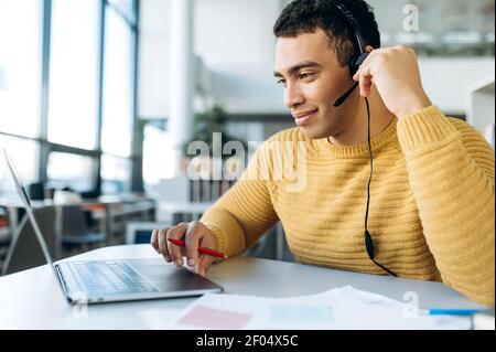 Nahaufnahme des freundlichen männlichen Betreibers des Callcenters am Arbeitsplatz. Attraktive Mitarbeiter, die einen Laptop verwenden und ein Headset tragen, beraten die Entfernung der Kunden Stockfoto
