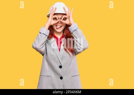 Junge Rotschopf Frau trägt Architekt Hardhut tun ok Geste wie Fernglas aufragend Zunge aus, Augen durch die Finger schauen. Verrückter Ausdruck. Stockfoto