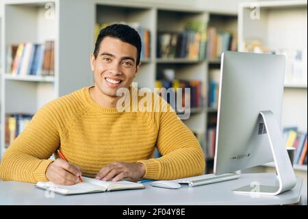 Erfolgreicher hispanischer männlicher Freiberufler am Arbeitsplatz, schaut lächelnd direkt auf die Kamera. Fröhlicher gutaussehender Kerl bei Webinar-Schulung oder Online-Konferenz Stockfoto