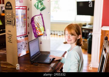 Ein junges, rot-köpfig Mädchen, das vor dem Computer-Homeschool-Setup im Esszimmer sitzt. Partition ist dekoriert und eingerichtet, um Ablenkungen zu reduzieren. Gir Stockfoto