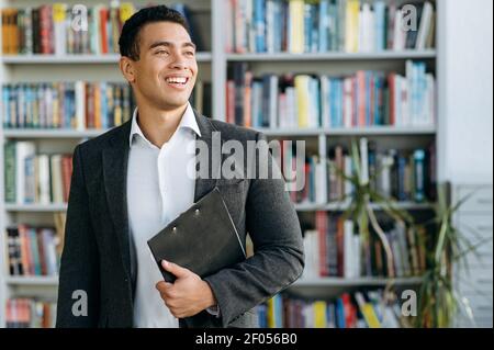 Zufriedener männlicher Manager steht im modernen Büro und lächelt. Erfolgreicher junger Geschäftsmann in stilvollem formellen Anzug, Blick auf die Seite, denken über neue Startup Stockfoto