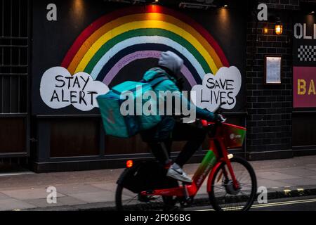 London, Großbritannien, 6. März 2021. Ein Lieferer fährt mit "Stay Alert" sein Fahrrad vor dem Regenbogen des NHS. Save Lives'-Zeichen während einer laufenden dritten Coronavirus-Sperre. Der Premierminister Boris Johnson hat einen Fahrplan zur Lockerung der Beschränkungen aufgestellt. Quelle: Dominika Zarzycka/Alamy Live News Stockfoto